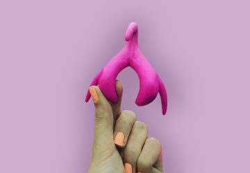 Hand holding a pink anatomical clitoris model on a purple background, wellness education