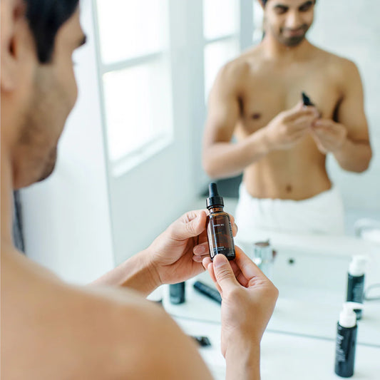 Shirtless man in bathroom applying beard oil with dropper in front of mirror
