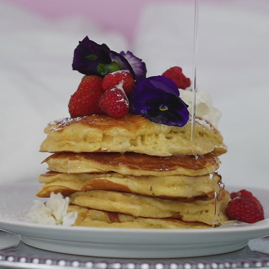 Stack of pancakes with syrup, berries, edible flowers, and whipped cream on white plate